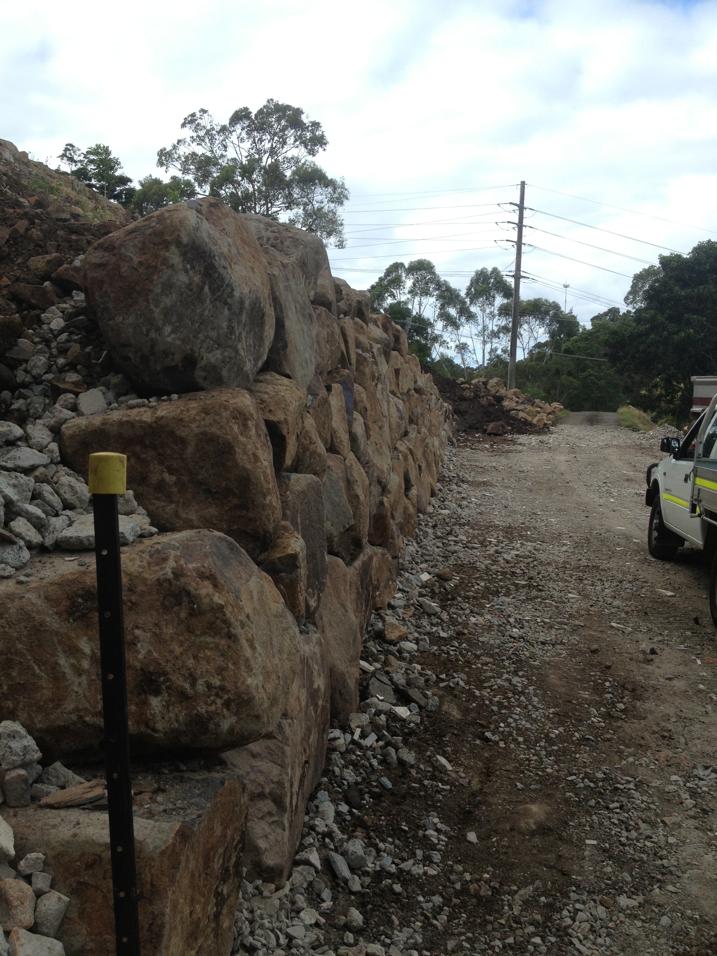 Large boulder retaining wall beside road with power lines and gravel