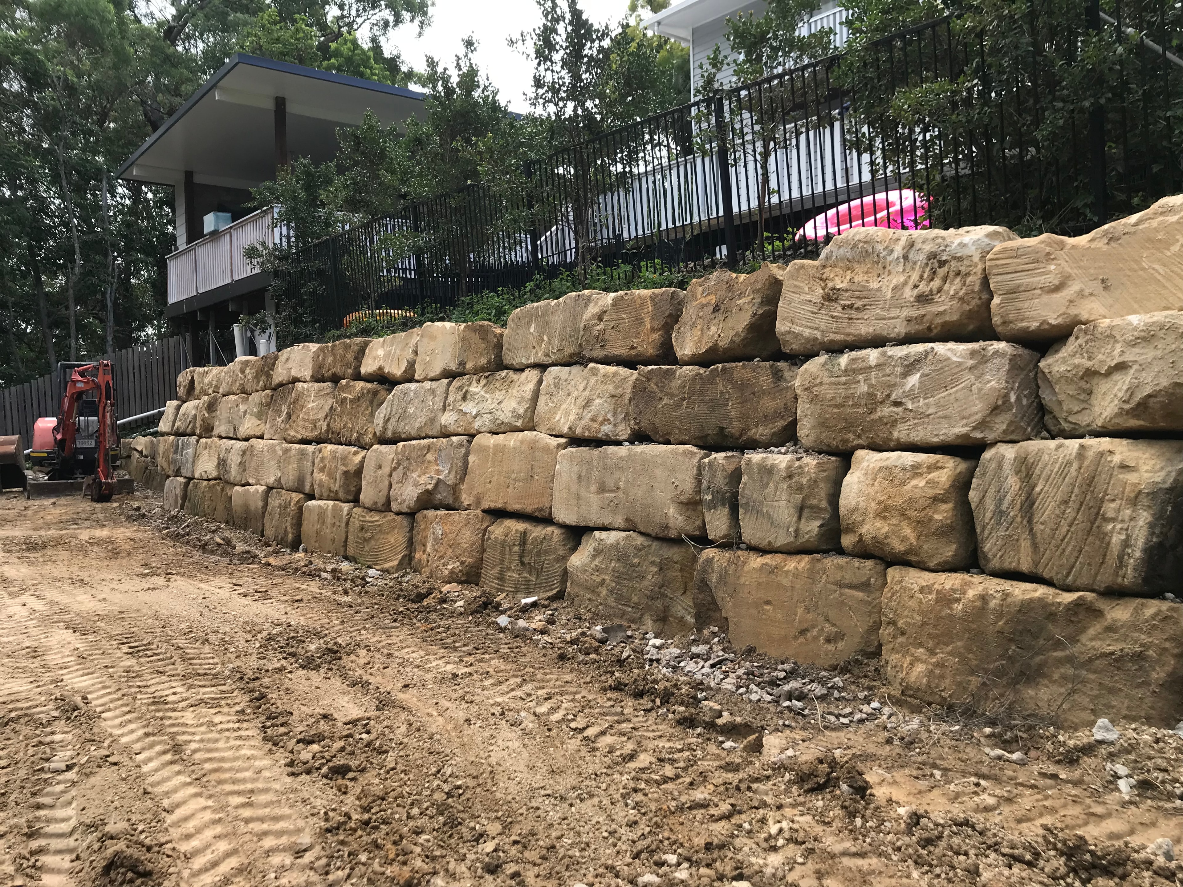 Sandstone block retaining wall under construction with excavator beside house